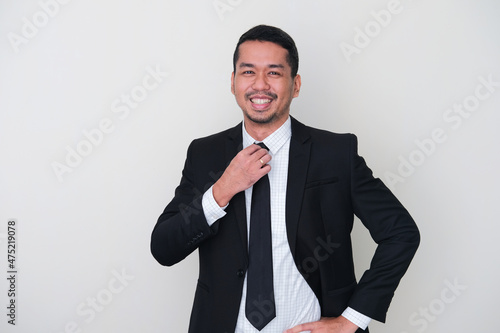 Adult Asian man wearing black suit showing high confidence with hand touching his tie photo