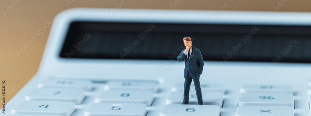 A miniature businessman figure wearing a suit while standing on top of a white calculator A miniature businessman figure wearing a suit while standing on top of a white calculator