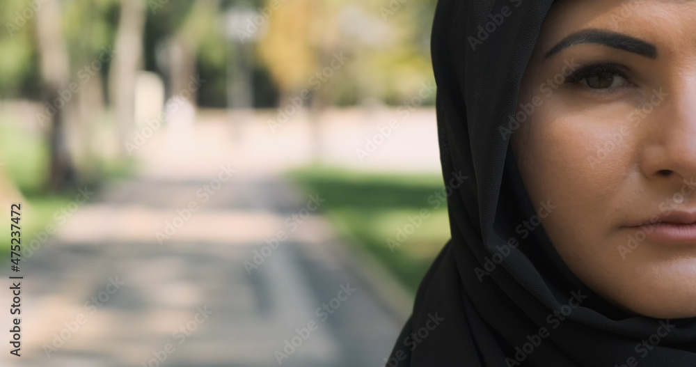 Half face portrait of muslim woman looking at camera, close-up. Arab woman pose in black hijab at city street. Serious, sad saudi female in traditional headscarf veil outdoors. With copy space text Half face portrait of muslim woman looking at camera, close-up. Arab woman pose in black hijab at city street. Serious, sad saudi female in traditional headscarf veil outdoors. With copy space text