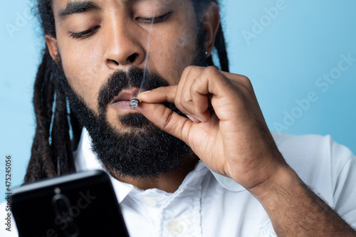 Black  man with dreadlocks and black beard smoking weed holding the cell blue bottom,  banground ,Marijuana joint in the hand, Concepts of medical marijuana use and legalization of the cannabis. tecno photo