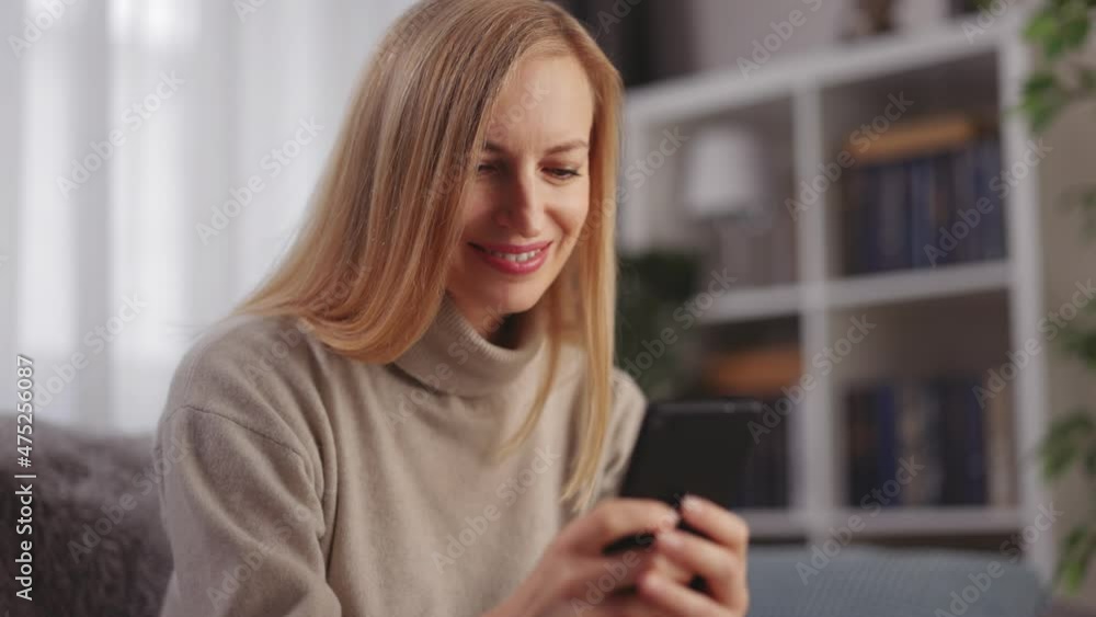 Positive caucasian woman typing messages on modern smartphone while resting on comfy couch. Online communication during free time at modern apartment. Positive caucasian woman typing messages on modern smartphone while resting on comfy couch. Online communication during free time at modern apartment.
