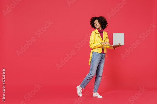 Full size body length bright happy fancy young curly black latin woman 20s wears yellow jacket go move hold use work on laptop pc computer look back isolated on plain red background studio portrait.
