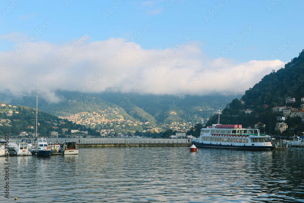 Panoramic view of lake with hills and morning clouds having green nature look and boats parked in small port in northern Italy 