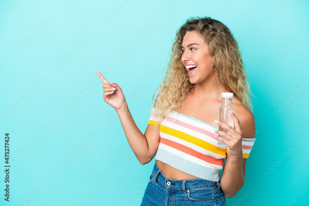 Young blonde woman with a bottle of water isolated on blue background pointing finger to the side and presenting a product Young blonde woman with a bottle of water isolated on blue background pointing finger to the side and presenting a product