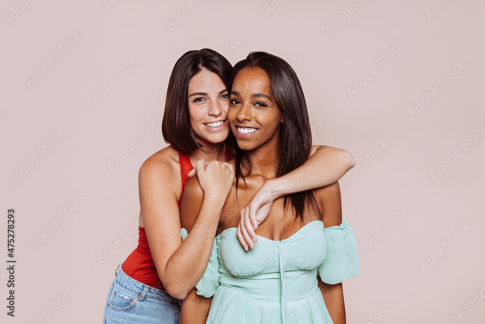 Mixed race and caucasian woman hugging at studio. Two cheerful multiracial female friends posing and looking at camera over beige background. Happy lifestyle concept. Mixed race and caucasian woman hugging at studio. Two cheerful multiracial female friends posing and looking at camera over beige background. Happy lifestyle concept.