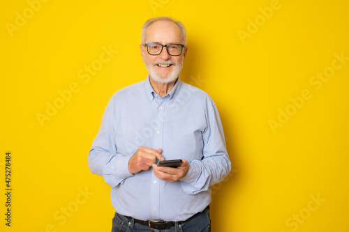 Portrait of happy european senior man using mobile phone and smiling isolated over yellow background.