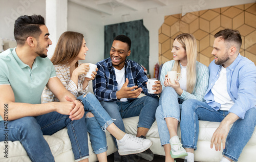Group of cheerful multiethnic friends having party at home, sitting on couch, drinking coffee