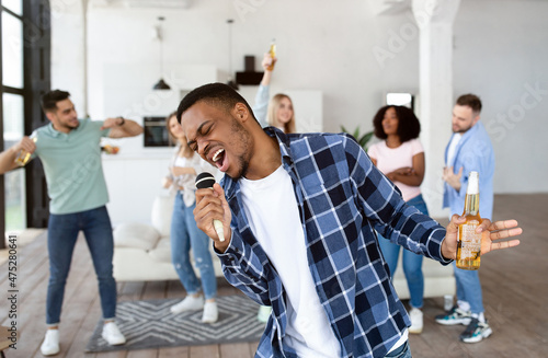 African American guy with beer bottle singing karaoke on home party with friends, singing songs into microphone