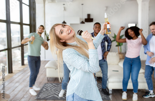 Positive millennial blonde lady with bottle of beer singing karaoke while having party with her friends at home