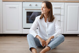 Indoor shot of smiling pensive woman wearing white shirt and jeans sitting on floor in kitchen and looking away with charming smile, posing against white kitchen set, expressing positive emotions.