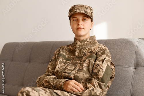 Indoor shot of serious young adult soldier woman wearing camouflage uniform sitting on cough and looking at camera with calm but satisfied facial expression.