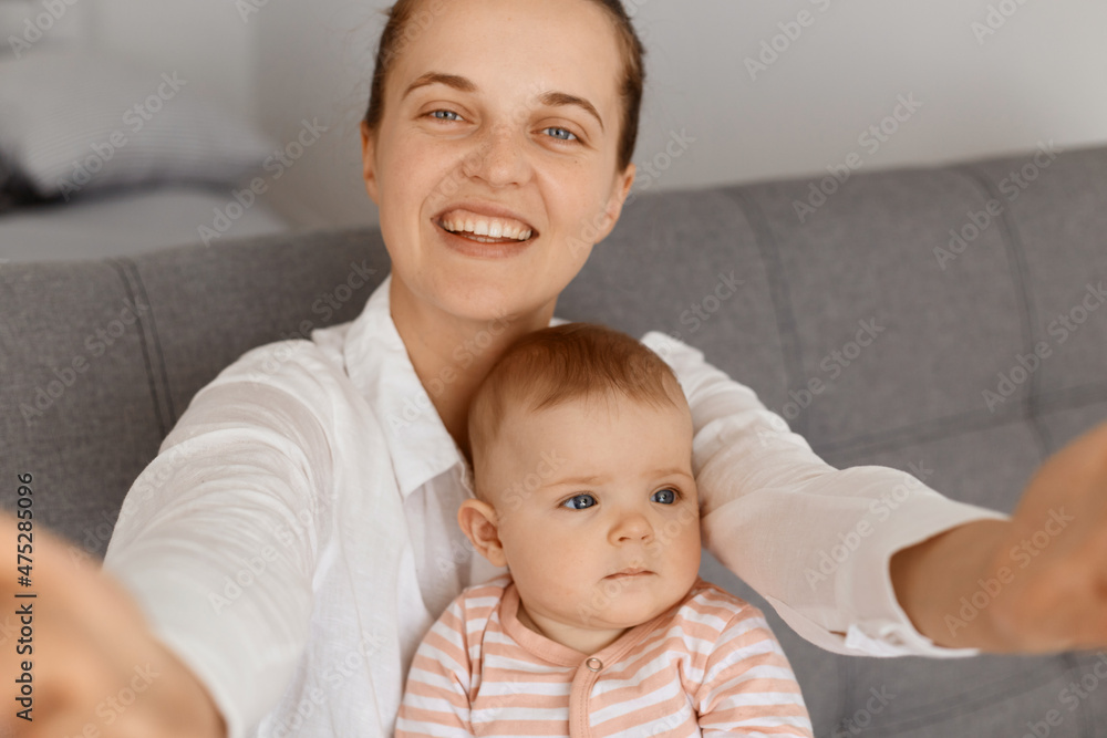 Smiling woman making selfie with her charming infant daughter POV, looking at camera with happy facial expression, point of view photo, female wearing white shirt. Smiling woman making selfie with her charming infant daughter POV, looking at camera with happy facial expression, point of view photo, female wearing white shirt.