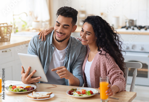 Cheerful Middle Eastern Spouses Using Digital Tablet In Kitchen While Eating Breakfast