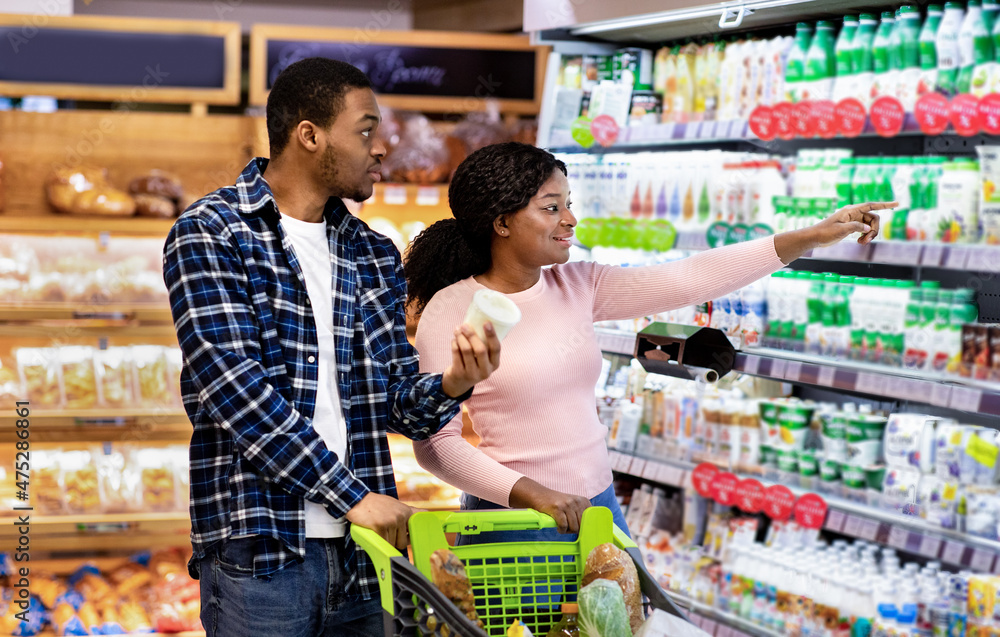 Young black couple with trolley full of products choosing diary products at huge supermarket