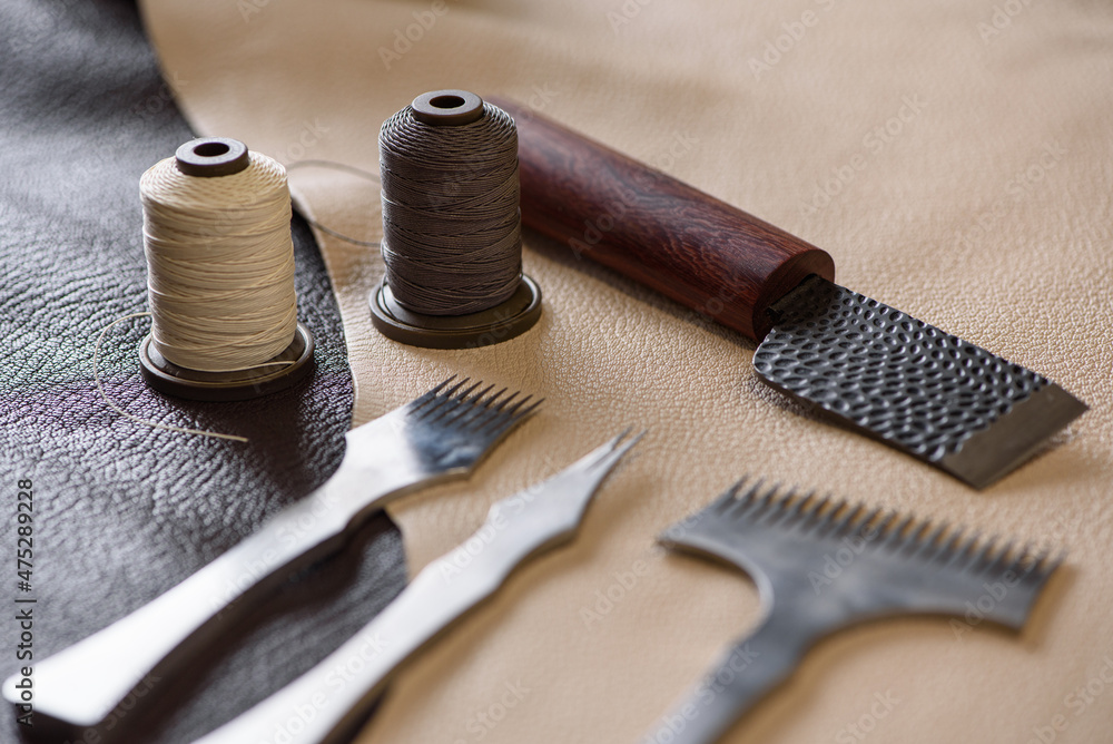 Shoemaker's tools laying on piece of beige and brown genuine leather in workshop. Workplace of shoemaker for sewing handmade shoes. Selective focus
