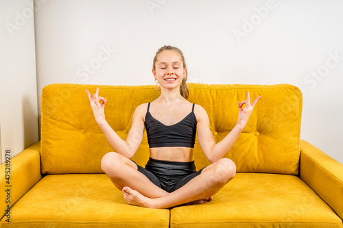 A little teenage girl is engaged in meditation and yoga on a yellow sofa, exercises at home, sitting in the lotus position