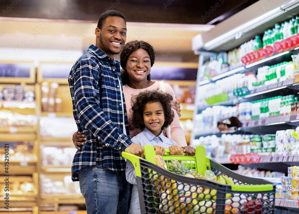 Portrait of joyful African American family with cute daughter posing and smiling at camera in big supermarket