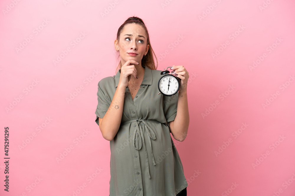 Fototapeta premium Young woman isolated on pink background pregnant and holding clock while thinking