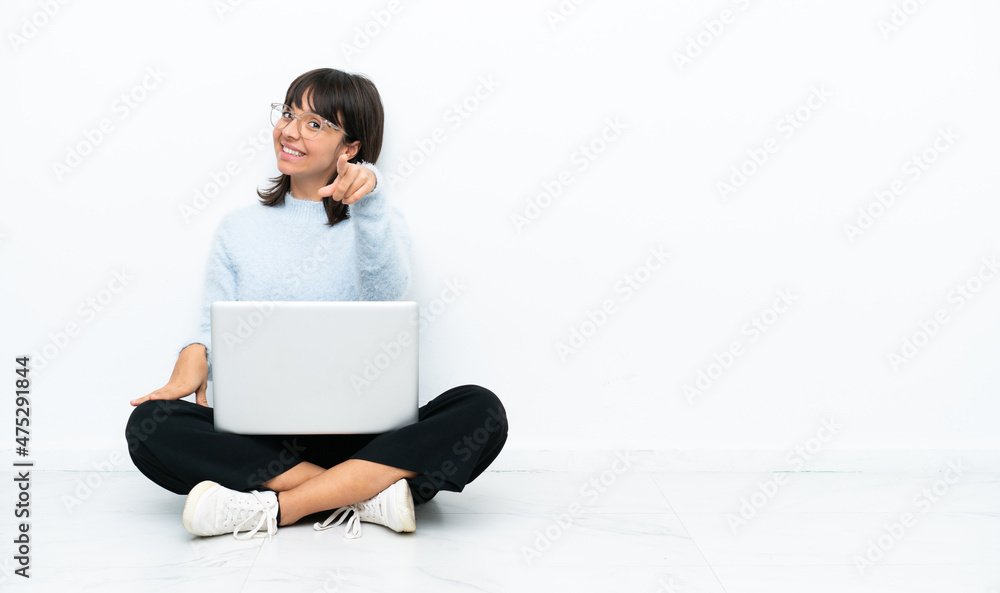 Young mixed race woman sitting on the floor with laptop isolated on white background points finger at you with a confident expression Young mixed race woman sitting on the floor with laptop isolated on white background points finger at you with a confident expression