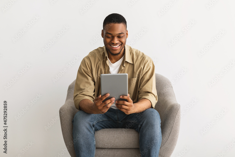 Black Guy Using Tablet Sitting In Chair Over Gray Background Black Guy Using Tablet Sitting In Chair Over Gray Background