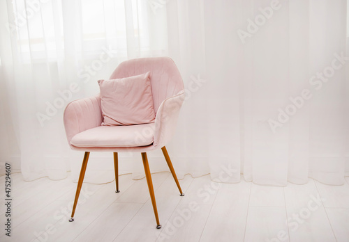 A light pink armchair stands by the window against a background of white airy curtains. A lonely chair in the room.