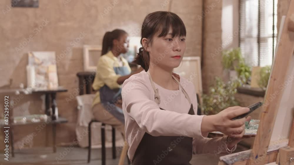 Waist-up of joyful Asian woman wearing apron, painting picture with putty knife on canvas in studio, then looking and smiling on camera Waist-up of joyful Asian woman wearing apron, painting picture with putty knife on canvas in studio, then looking and smiling on camera