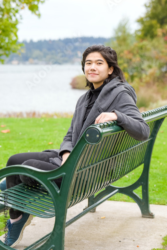 Biracial Asian teen girl sitting on park bench by lake