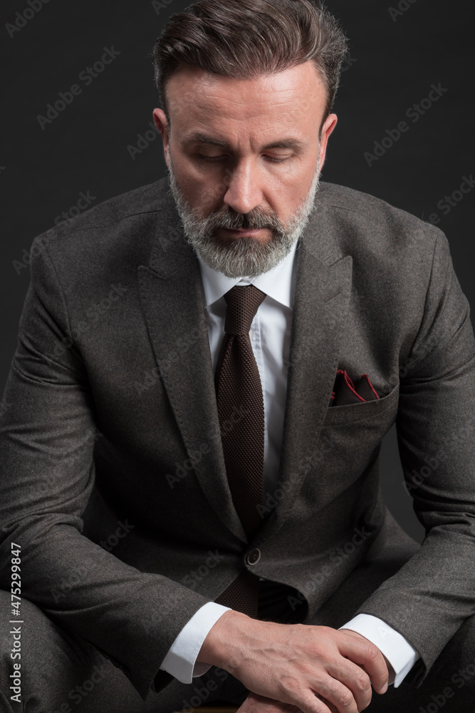 Portrait of adult businessman wearing trendy suit and sitting in modern studio on stylish chair against the black background