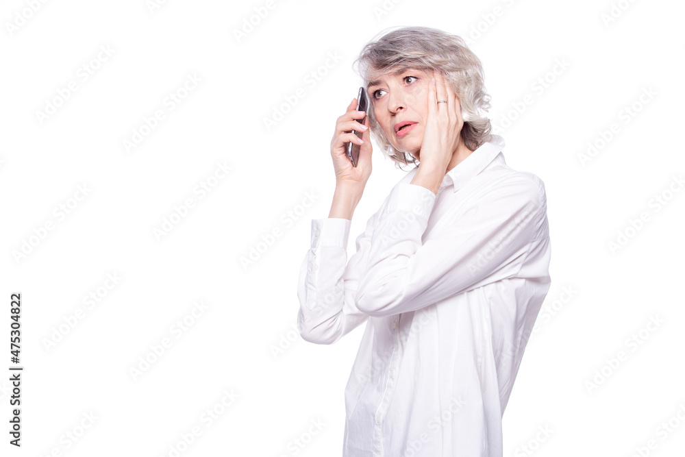 An adult gray-haired woman looks scared and upset, holding her head while talking on a mobile phone isolated on white background. Phone fraud An adult gray-haired woman looks scared and upset, holding her head while talking on a mobile phone isolated on white background. Phone fraud