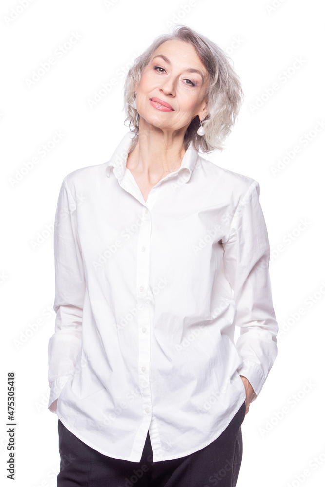 Happy trendy senior woman with a beautiful smile wearing a fashionable shirt standing with her hands in her pockets looking at the camera isolated on white background Happy trendy senior woman with a beautiful smile wearing a fashionable shirt standing with her hands in her pockets looking at the camera isolated on white background