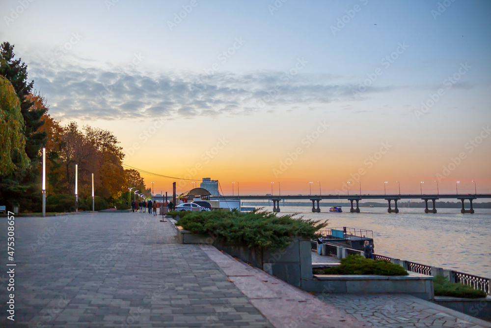 Fototapeta premium River embankment in the evening, sky and bridge in the distance, empty sidewalk in the foreground for juices space text, blurred background