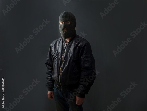 Fototapete a man stands against a dark background in a black bomber jacket and a balaclava