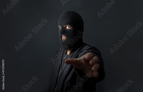Fototapeta a man stands against a dark background in a black bomber jacket and a balaclava mask on his face and pulls his hand into the camera