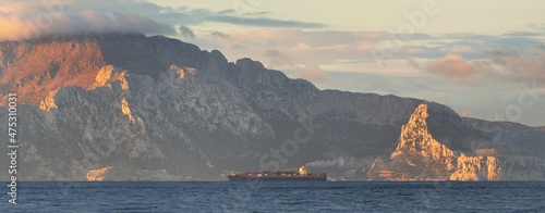 Cargo ship sailing near the Rock of Gibraltar at sunrise, a view from the yacht. British Overseas Territory. Epic cloudscape. Travel destinations, logistics, global communications photo