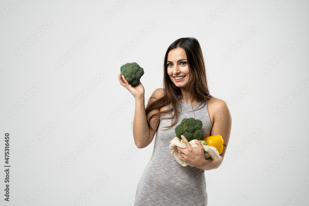 Young brunette girl holds vegetables in mesh eco bag in one hand and a broccoli in another. Zero waste concept, plastic-free, eco-friendly shopping, vegan. Young brunette girl holds vegetables in mesh eco bag in one hand and a broccoli in another. Zero waste concept, plastic-free, eco-friendly shopping, vegan.