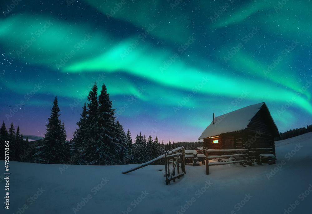 Fantastic winter landscape with wooden house with light in window in snowy mountains and northen light in night sky. Christmas holiday and winter vacations concept