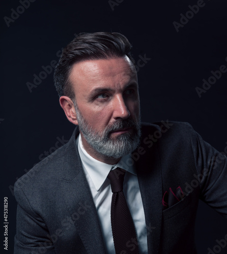 Portrait of a stylish elegant senior businessman with a beard and casual business clothes in photo studio isolated on dark background gesturing with hands