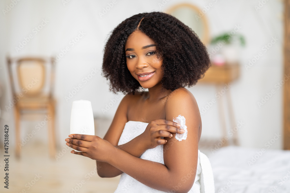 Millennial black lady applying cream, lotion or body milk after bath, making domestic beauty procedure indoors Millennial black lady applying cream, lotion or body milk after bath, making domestic beauty procedure indoors