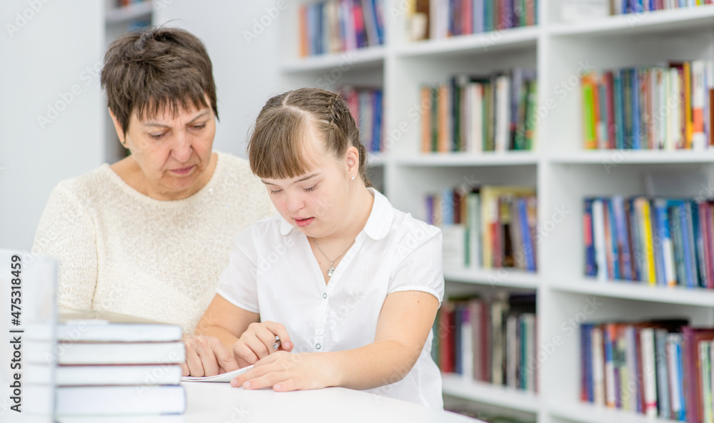Senior teacher and girl with Downs syndrome doing homework at library. Education for disabled children concept. Empty space for text Senior teacher and girl with Downs syndrome doing homework at library. Education for disabled children concept. Empty space for text