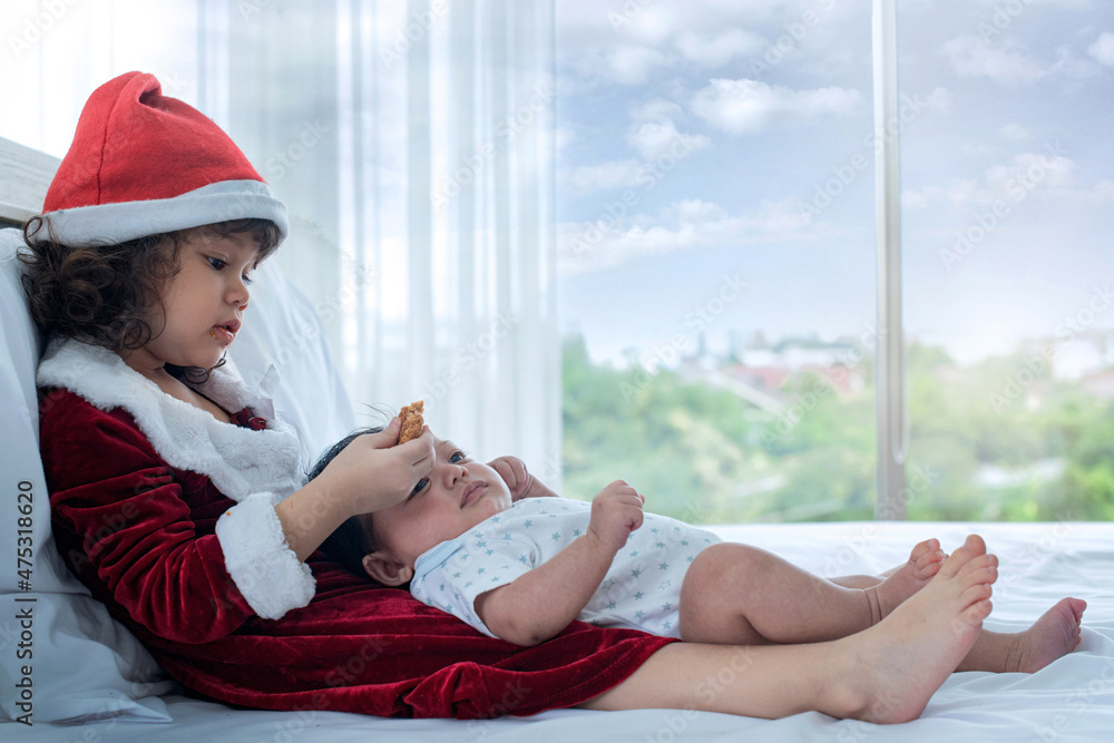 Cute little girl in Santa costume takes care of her sister with love while enjoying cookies in hand, baby lying on her lap in bedroom  near window at home