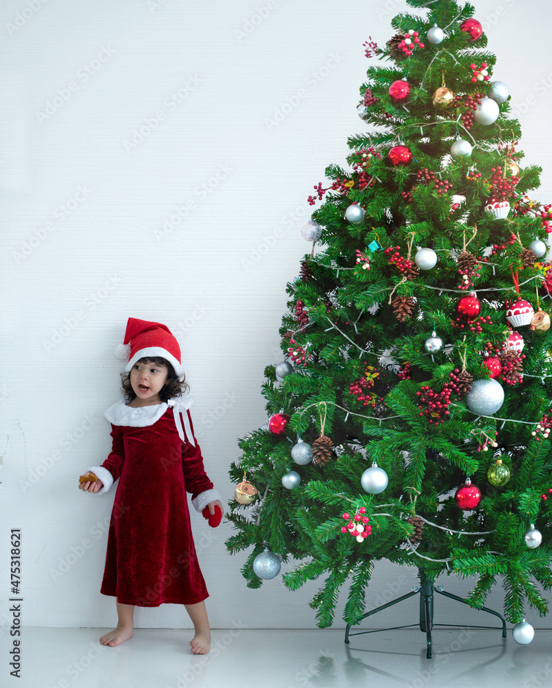 Cute little girl in Santa costume standing near her Christmas tree on white wall, Christmas Concept