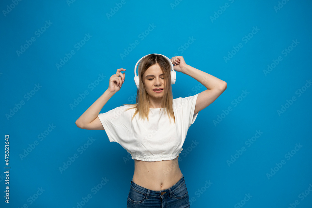 Beautiful attractive young blond woman wearing white t-shirt and glasses in white headphones listening music and dancing on blue background in studio. Relaxing and enjoying. Lifestyle. Beautiful attractive young blond woman wearing white t-shirt and glasses in white headphones listening music and dancing on blue background in studio. Relaxing and enjoying. Lifestyle.
