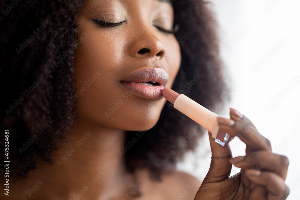 Obraz premium Closeup portrait of young black lady applying lipstick with closed eyes, taking care of her appearance indoors
