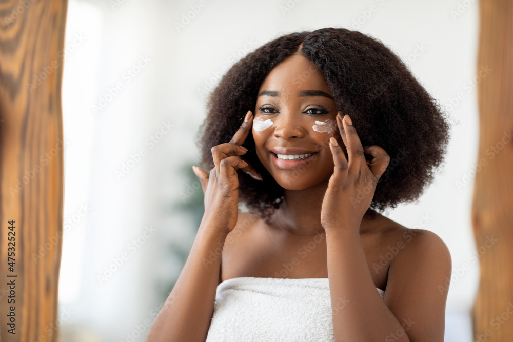 Attractive millennial black woman applying face cream after shower near mirror indoors. Spa and wellness concept