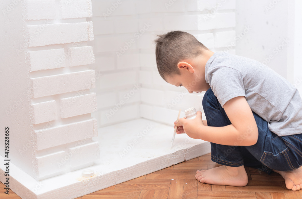 Child boy sits on the floor and paints a decorative fireplace with white paint gouache. Home improvement, DIY concept. Selective focus Child boy sits on the floor and paints a decorative fireplace with white paint gouache. Home improvement, DIY concept. Selective focus