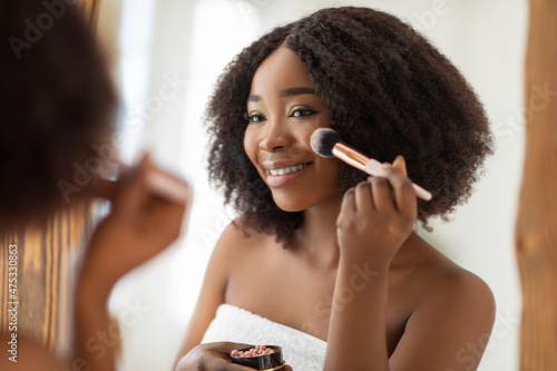Natural makeup for young skin. Sexy black lady putting on blusher or powder with brush in front of mirror indoors