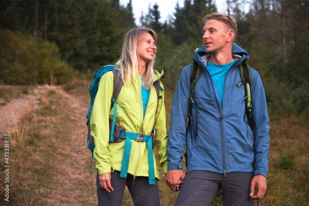 Happy loving couple holding hands and smiling while hiking together in mountain forest. Woman and man travelers wearing sports jackets and carrying backpacks while standing on grassy forest road.