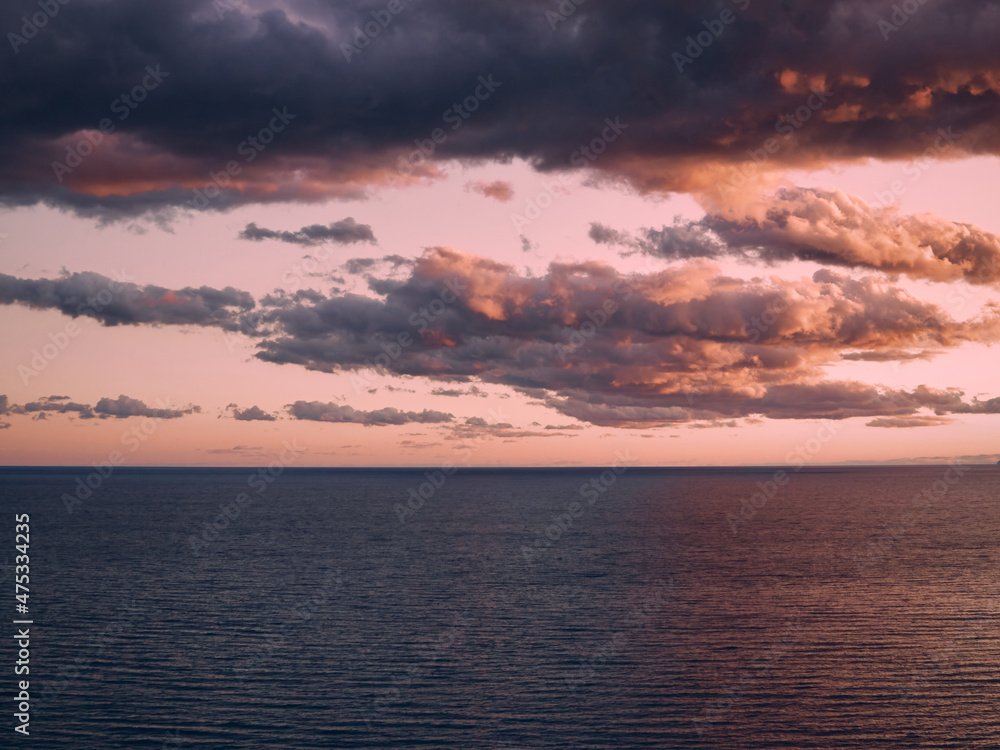 There are light ripples on the surface of the sea, the horizon is clearly visible, the rays of the setting sun illuminate the sky and the sea, clouds in the sky