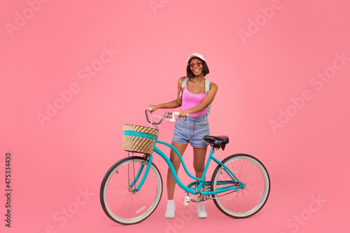 Positive young black lady in fashionable summer clothes posing with classic bicycle on pink studio background © Prostock-studio