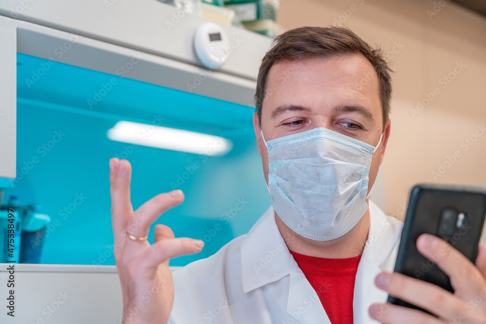 Fototapeta premium Excited scientist wearing white coat showing emotions of joy about victory in online lottery in room with laboratory box in UV ligh. Amazed male in medical mask win money celebration success